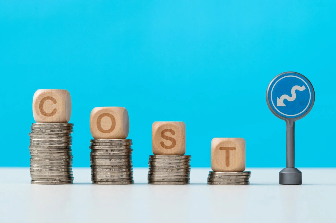 Wooden blocks spelling 'COST' on descending coin stacks with a blue winding arrow sign on a blue background.